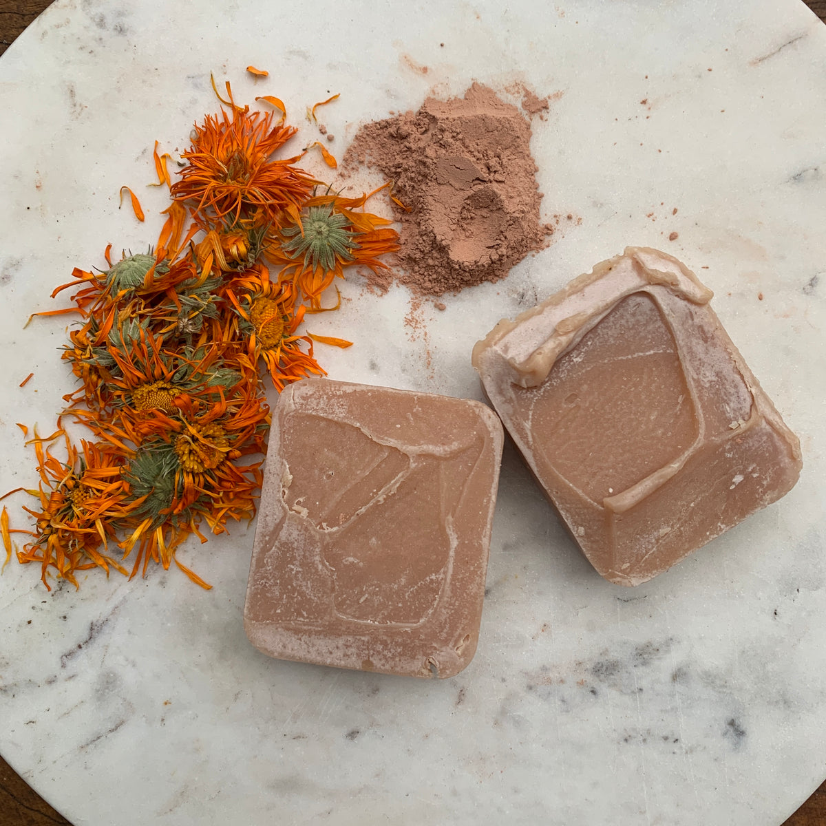 Two bars of pink clay soaps, next to dried pink clay and calendula flowers on a marble surface The Herbal Gardener