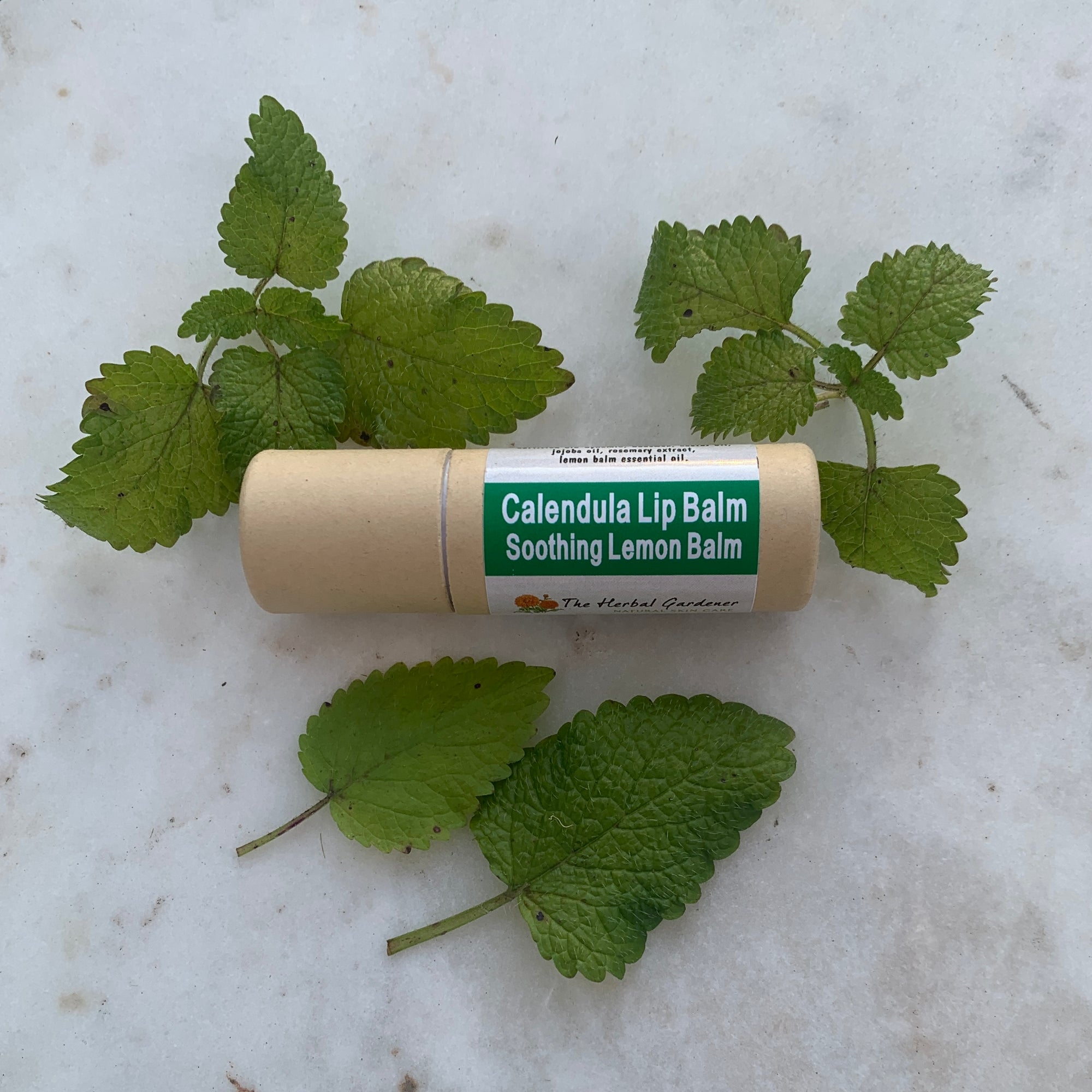 One calendula lip balm labeled 'Soothing Lemon Balm' placed on a surface with natural lemon balm leaves around it.