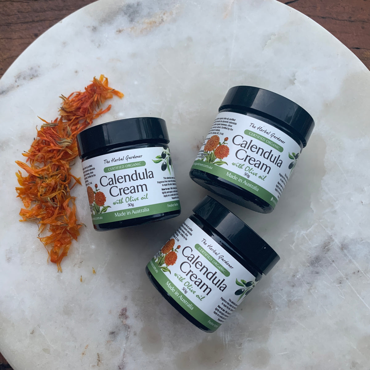 Calendula Cream Three Pack 50gr each placed on a marble surface next to dried calendula flowers The Herbal Gardener