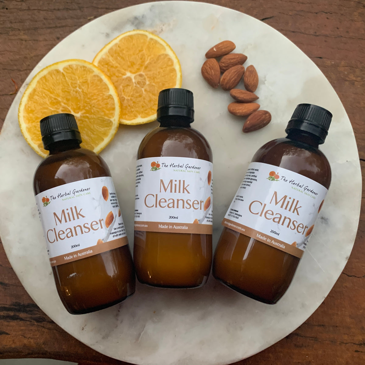 Three bottles of 200 mL Milk Cleanser on a marble and wooden background with sliced oranges and almonds