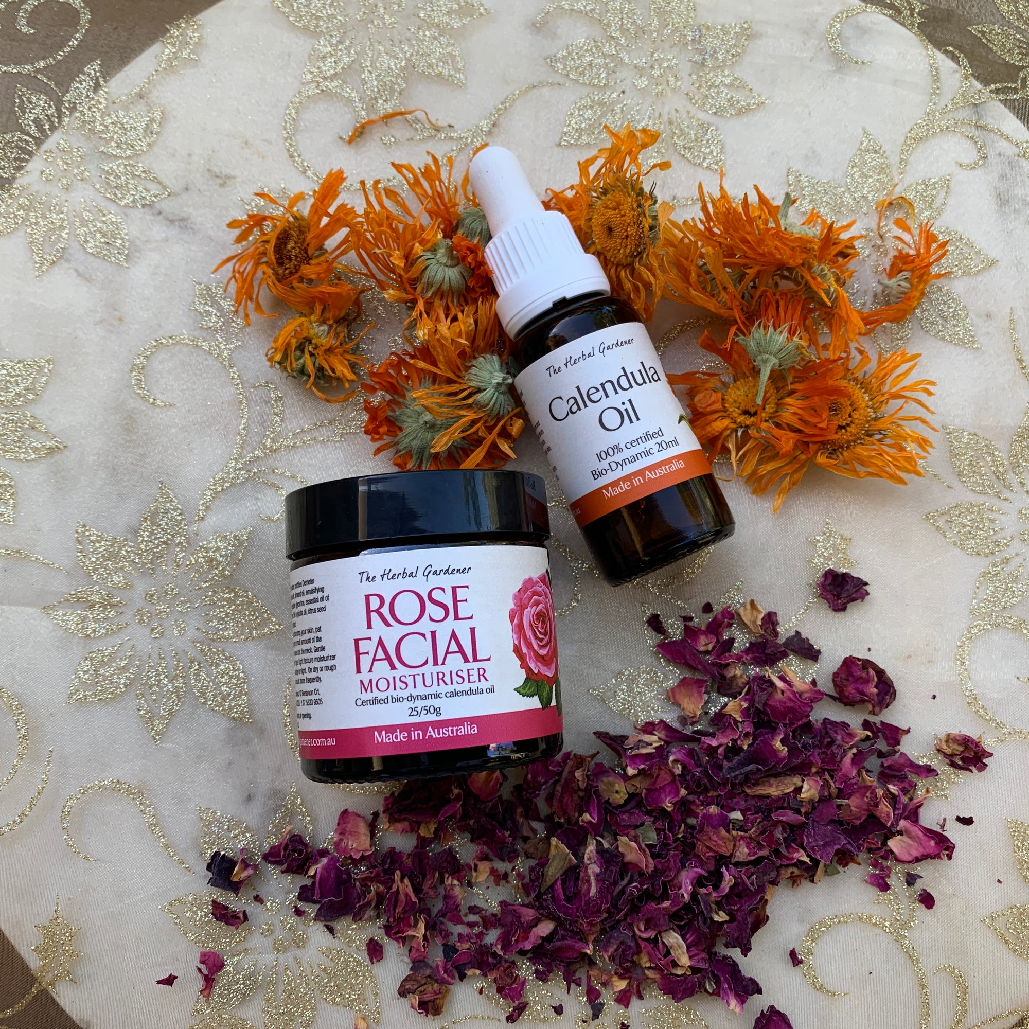Glowing Rose and Calendula Pack including the rose facial moisturiser 25gr and calendula oil 20mL on a sparkly fabric with dried rose and calendula The Herbal Gardener