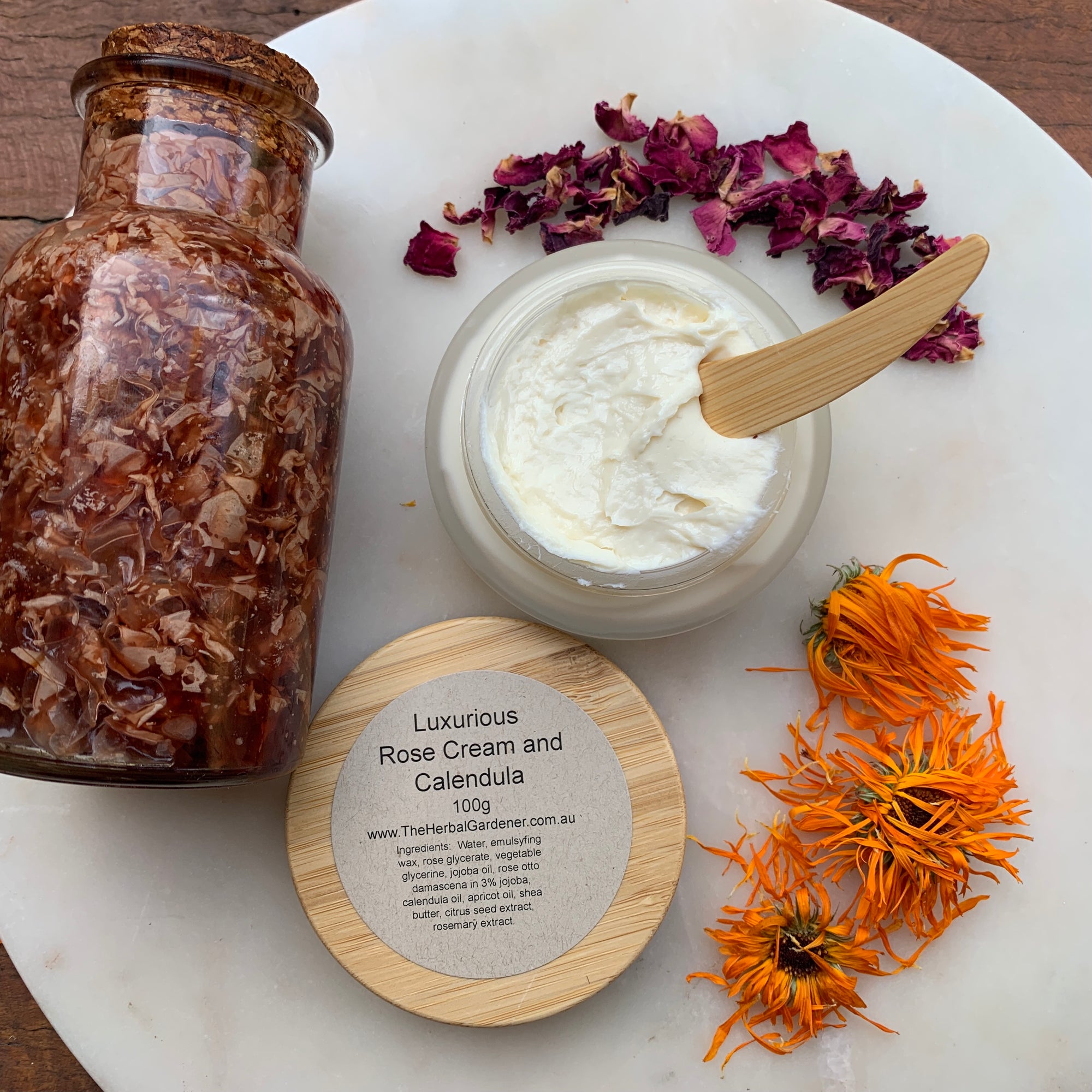 Luxurious rose and calendula cream 100gr with a bamboo lid, alongside a wooden spoon, dried rose petals, and fresh calendula flowers The Herbal Gardener