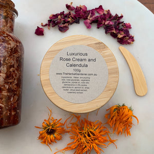 Luxurious rose and calendula cream 100gr with a bamboo lid, alongside a wooden spoon, dried rose petals, and fresh calendula flowers The Herbal Gardener