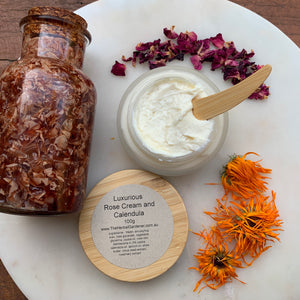 Luxurious rose and calendula cream 100gr with a bamboo lid, alongside a wooden spoon, dried rose petals, and fresh calendula flowers The Herbal Gardener